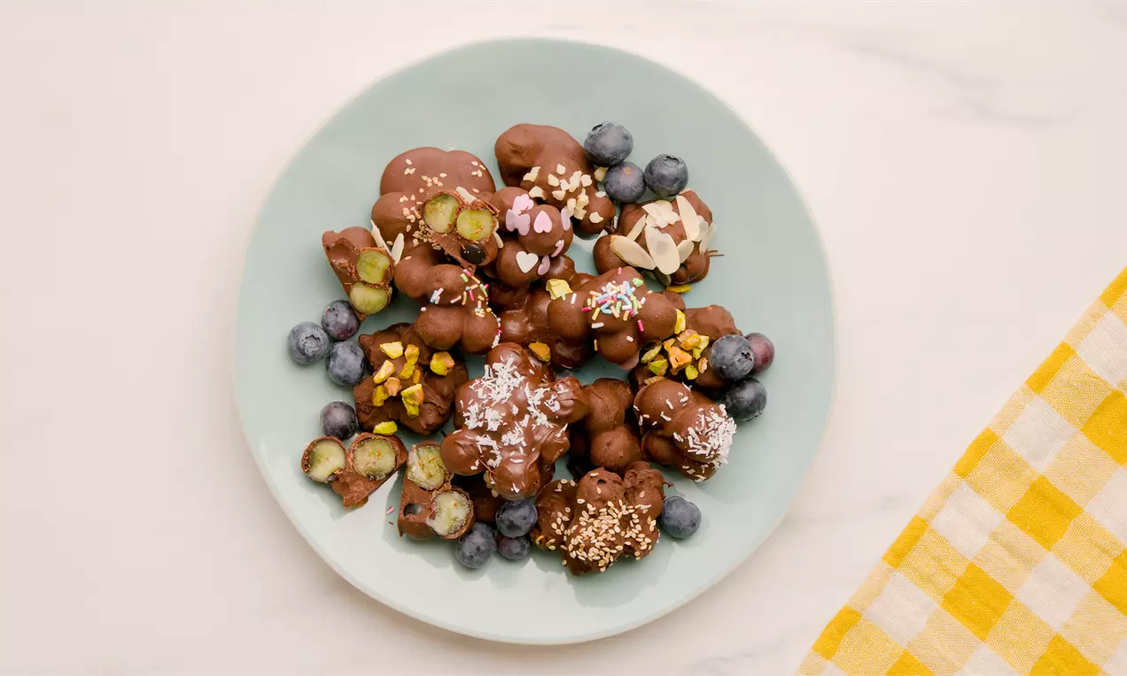 Rocas de arándanos cubiertas de chocolate suave con decoraciones de frutos secos, coco rallado y caramelos en un plato verde pastel