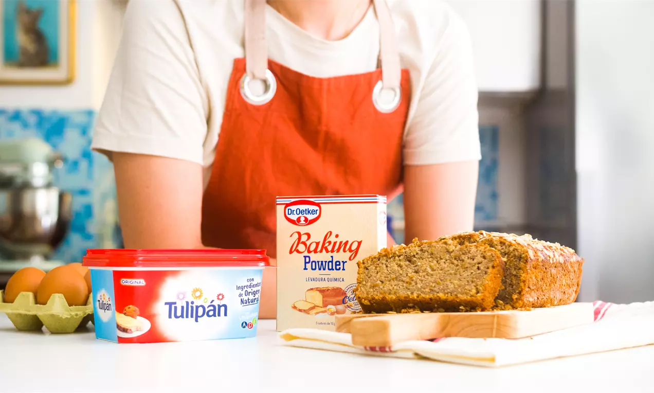 Bizcocho rectangular de tono dorado con trozos de avena en la superficie sobre una tabla de madera junto a ingredientes dispersos en una cocina