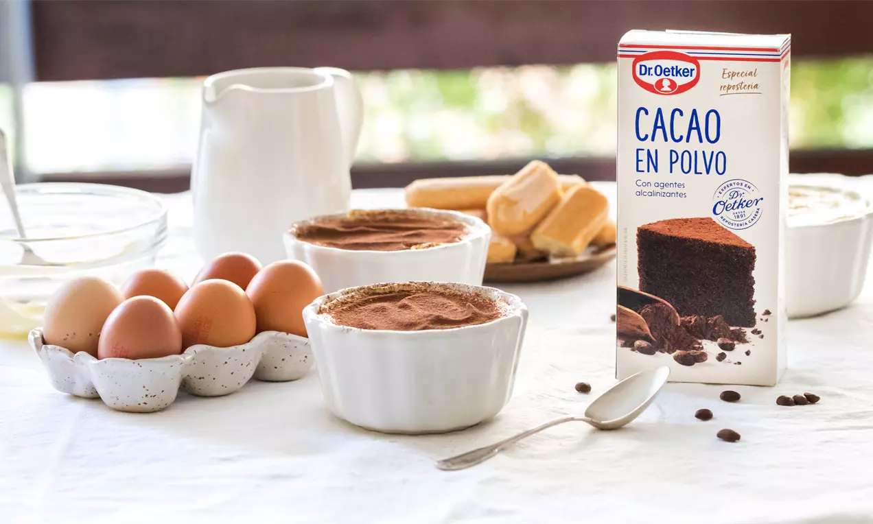 Tiramisú en recipientes cerámicos blancos con capa superior de cacao marrón suave, rodeado de huevos, bizcochos y crema sobre un mantel blanco