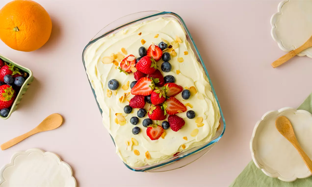 Tiramisú en fuente rectangular de crema pálida cubierta con fresas, arándanos, frambuesas y almendras laminadas, rodeado de utensilios sobre un fondo claro