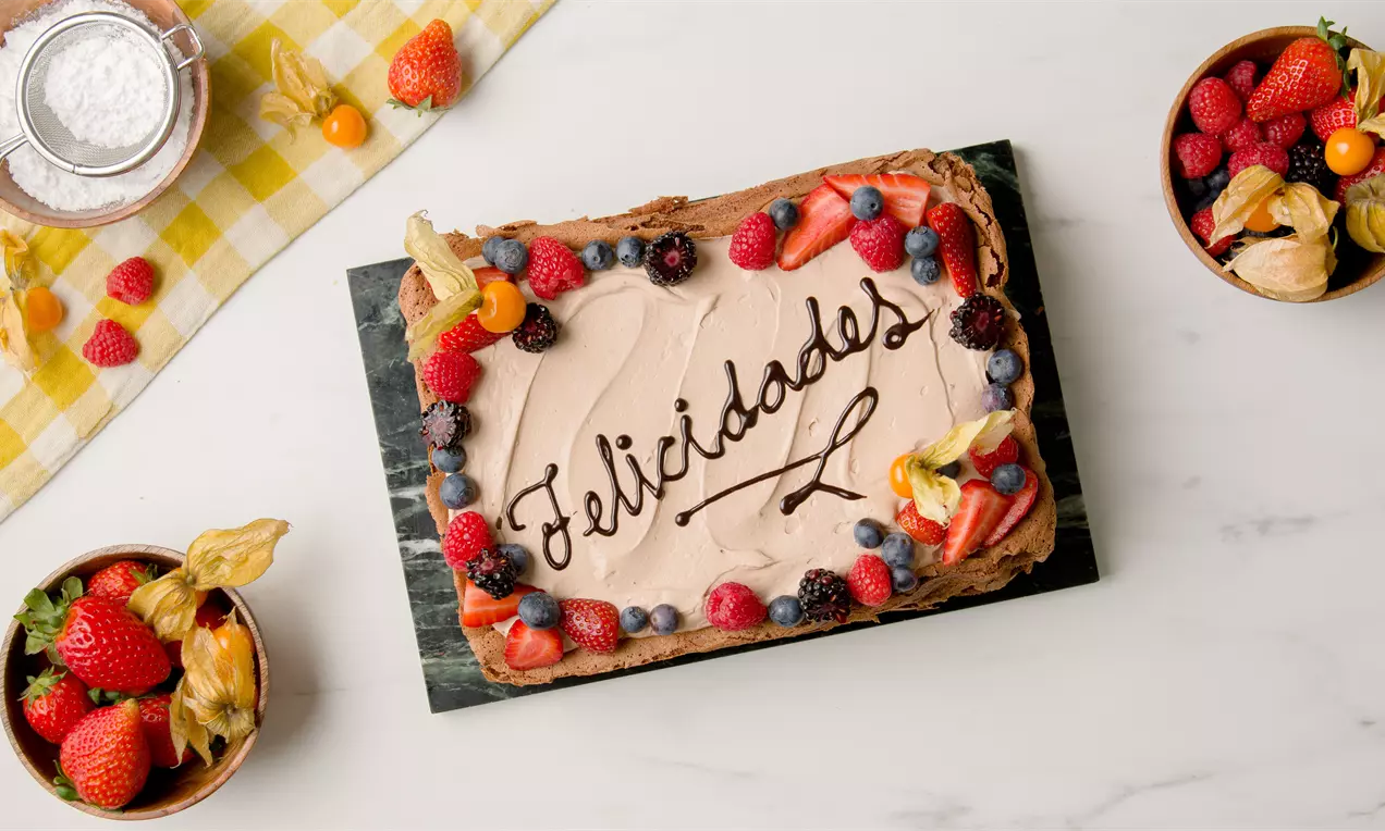 Tarta rectangular de chocolate con crema marrón clara, mensaje "Felicidades" en chocolate oscuro y bordes decorados con frutas frescas