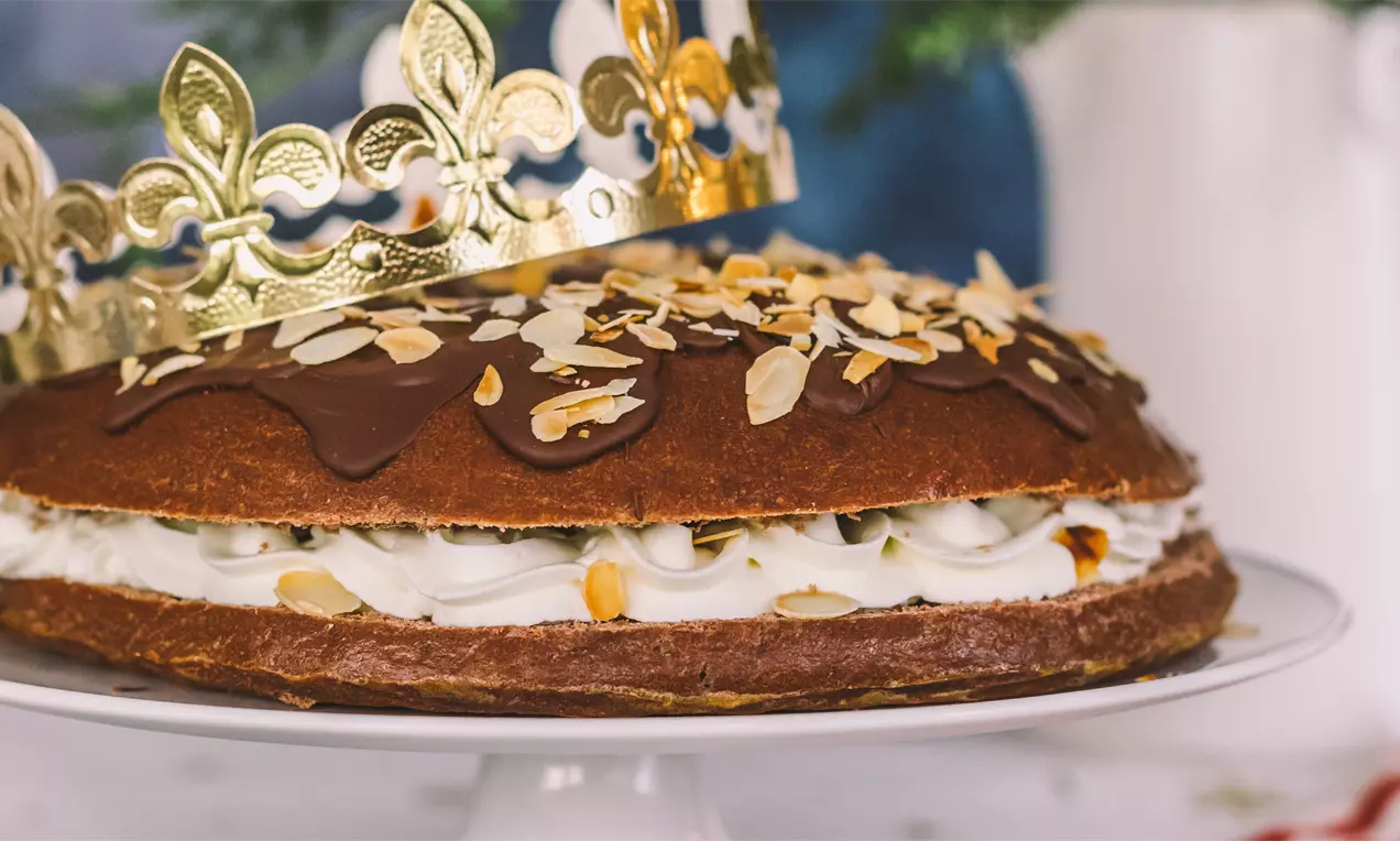 Roscón dorado con capa de chocolate brillante, almendras laminadas, relleno de nata blanca y corona dorada sobre un plato blanco