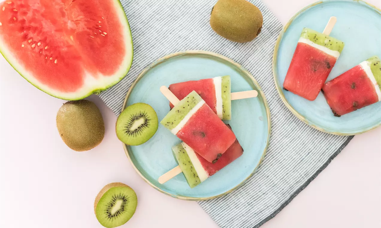 Polos rectangulares de sandía roja con capa de kiwi verde moteado y blanco, sobre platos azul claro con rodajas de kiwi y media sandía