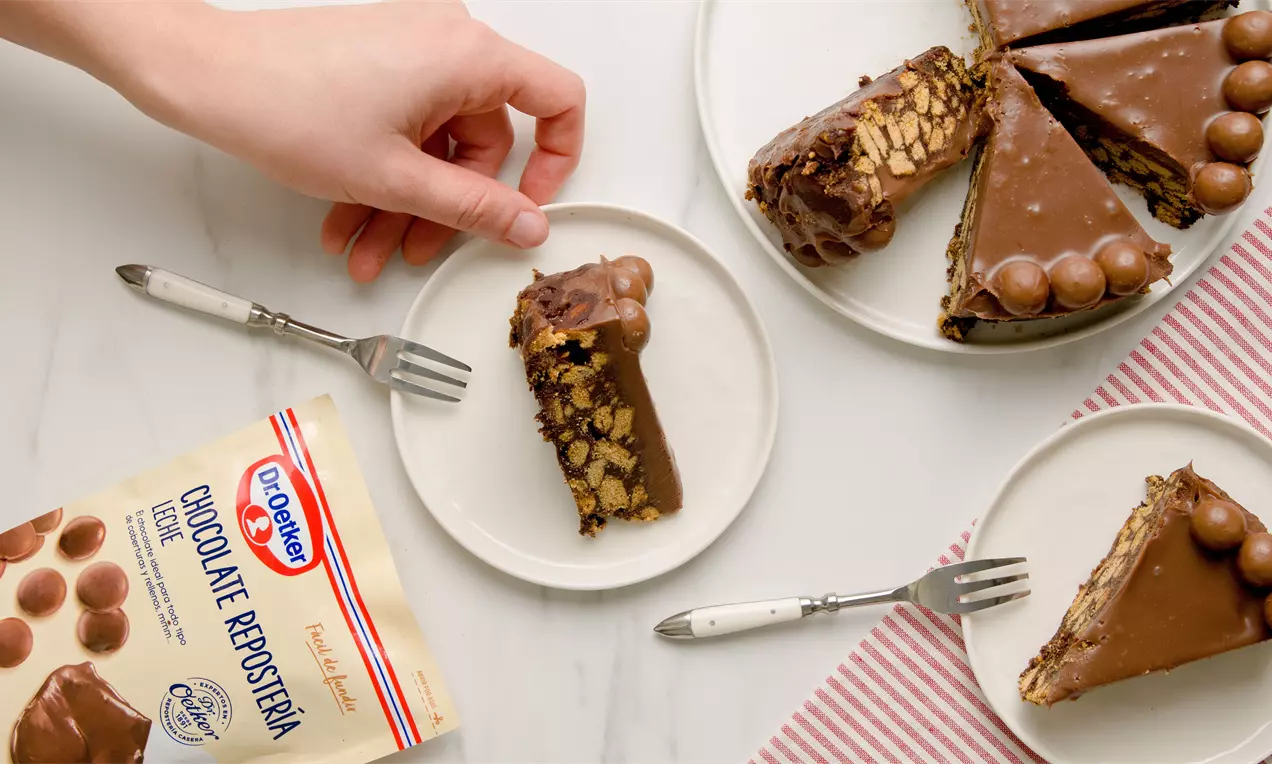 Tarta redonda de chocolate con cobertura brillante, capas de galletas y trozos en su interior, decorada con esferas en un plato blanco