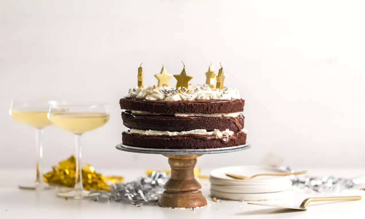 Pastel de capas de bizcocho de chocolate y crema blanca intercalada, decorado con estrellas doradas y velas sobre un soporte madera
