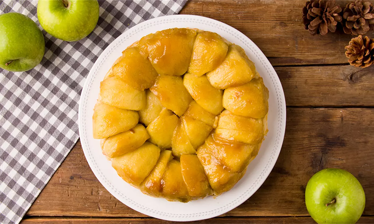 Tarta Tatín circular de manzanas doradas y brillantes acomodadas en espiral sobre un plato blanco, con manzanas verdes y piñas decorativas