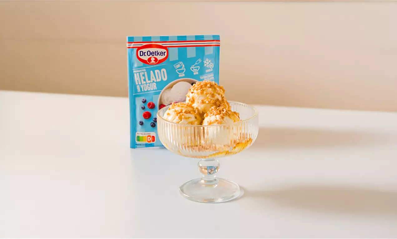 Helado cremoso color marfil con textura suave, adornado con miel y nueces troceadas servido en copa transparente sobre mesa blanca