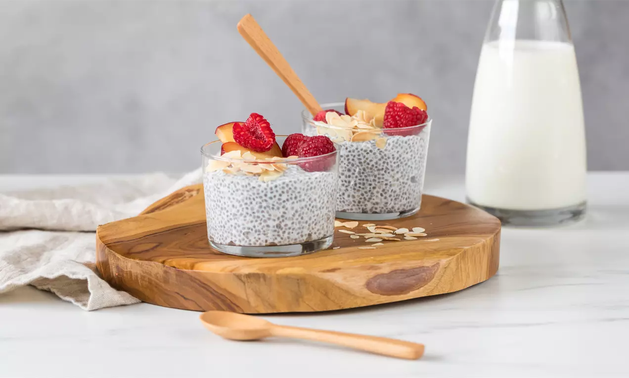 Pudin gris moteado de chía servido en vasos transparentes, decorado con frambuesas, rodajas de durazno y hojuelas de avena sobre base de madera
