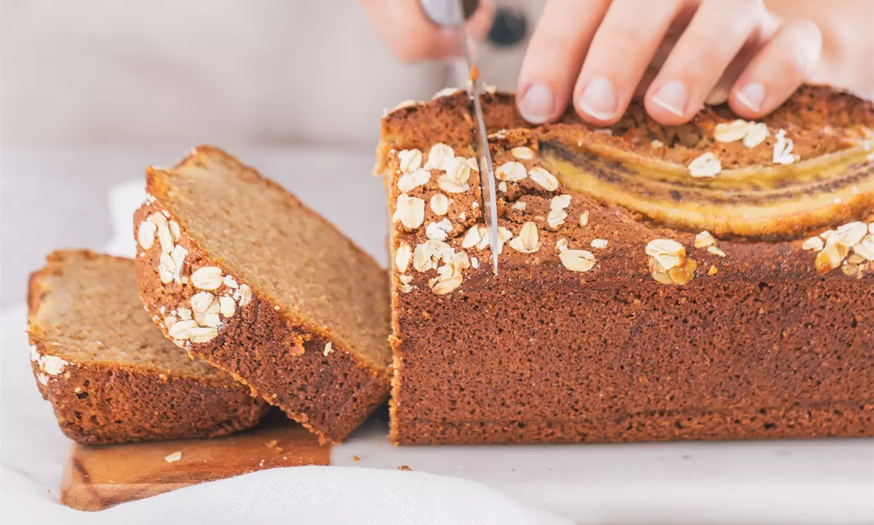Plum cake rectangular dorado con interior esponjoso y corteza decorada con rodajas de plátano y copos de avena sobre una tabla blanca