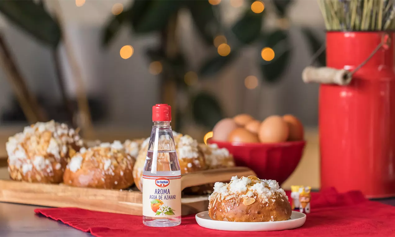 Mini roscón dorado y redondeado cubierto de azúcar granulada blanca sobre plato blanco, rodeado de ingredientes y decoración festiva