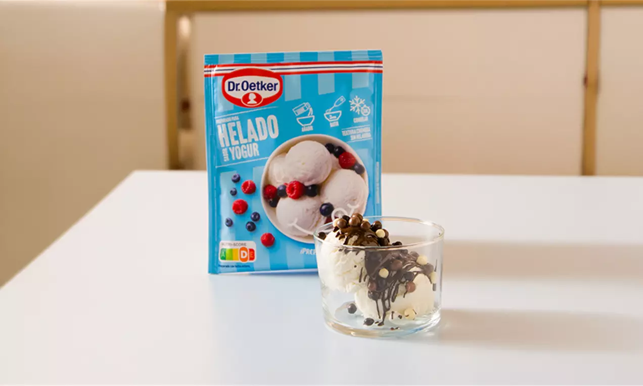 Bolitas de helado blanco cremoso en vaso transparente, decoradas con chocolate líquido y pequeños trozos crujientes sobre mesa blanca