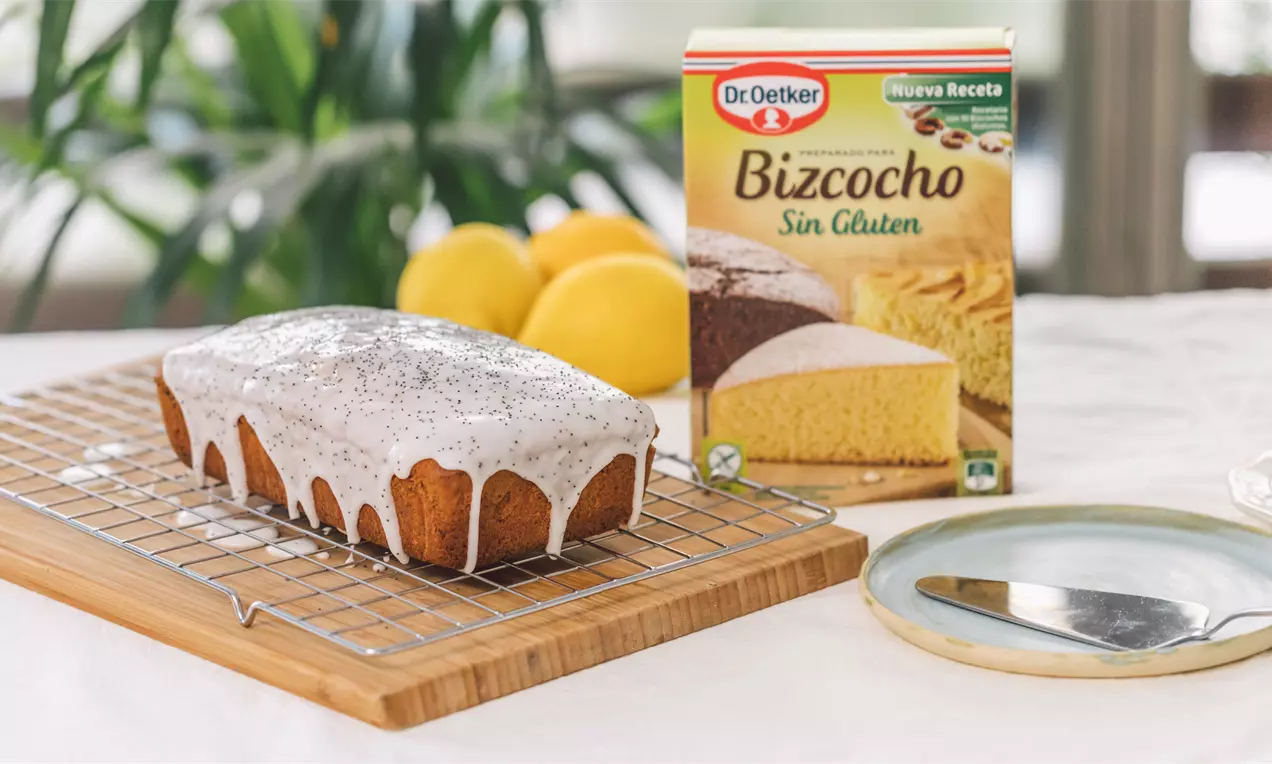 Bizcocho rectangular dorado con glaseado blanco brillante y semillas de amapola, sobre rejilla de madera con limones de fondo