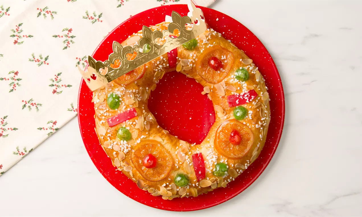 Roscón circular con masa dorada y suave, decorado con frutas glaseadas, rodajas de naranja, azúcar perlado y una corona dorada sobre un plato rojo