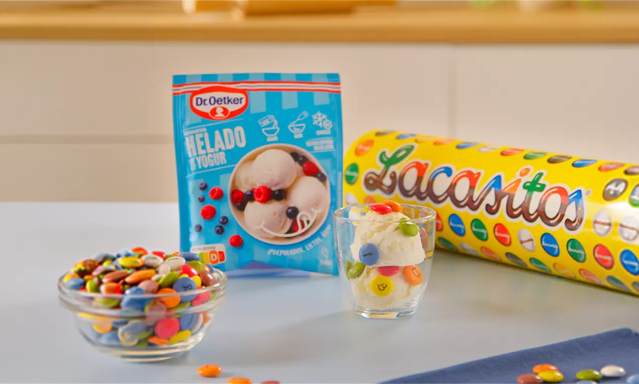 Helado blanco cremoso en vaso transparente decorado con Lacasitos coloridos y textura lisa, rodeado de tarros y envases coloridos sobre mesa clara