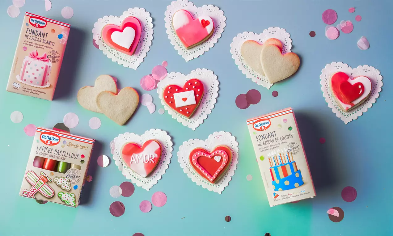 Galletas en forma de corazón decoradas con glaseado rojo, blanco y detalles románticos sobre blondas de papel y fondo azul con confeti