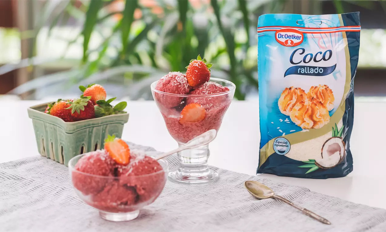Helado rosado y cremoso de fresas, coco y plátano en copas de cristal, decorado con fresas frescas sobre un mantel claro
