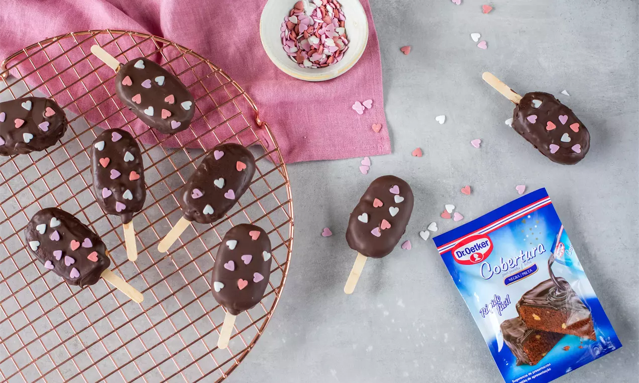 Cake pops alargados con cobertura de chocolate brillante decorados con pequeños corazones de azúcar rosa, blanco y lila sobre rejilla cobriza