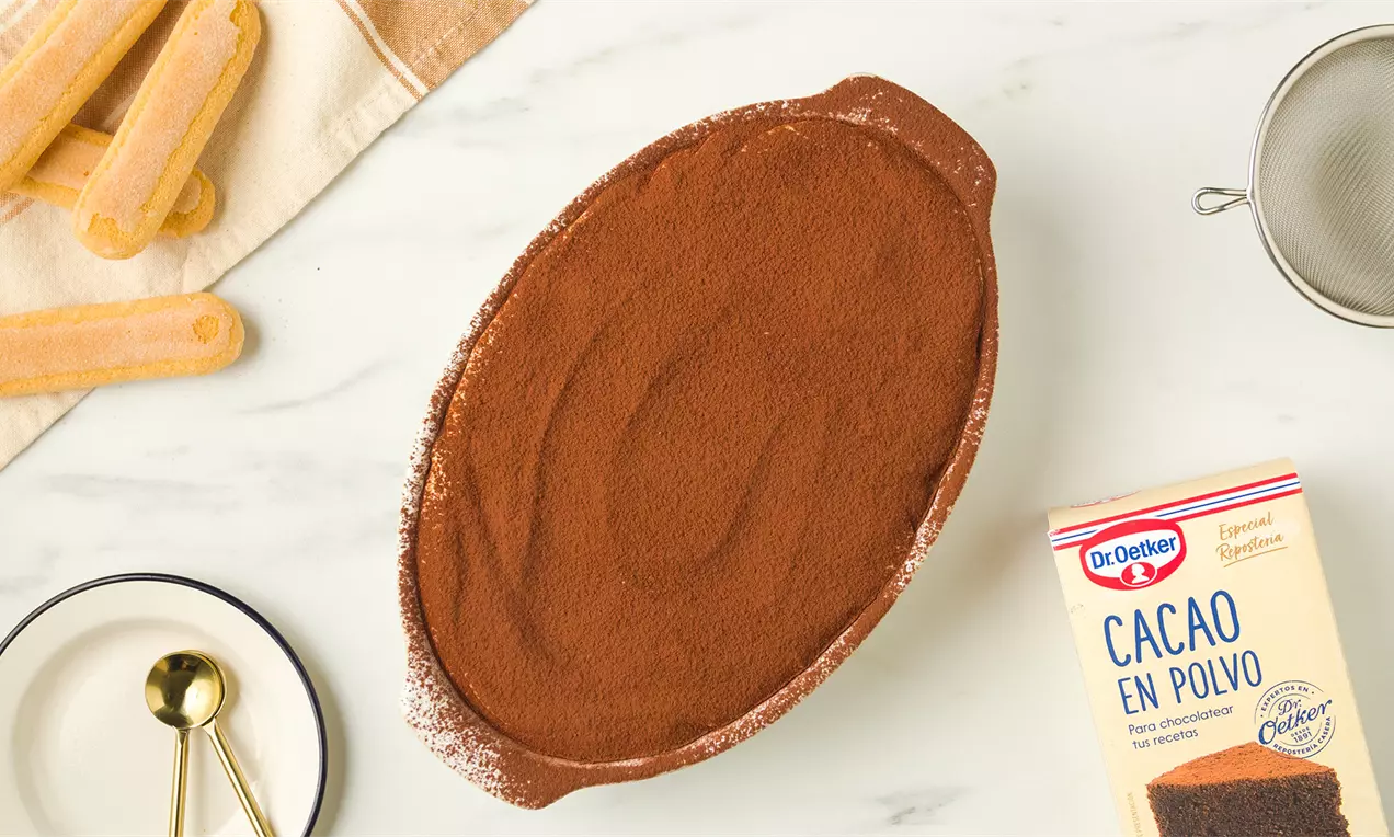 Tiramisú ovalado cubierto con una capa homogénea de cacao en polvo marrón sobre una fuente de cerámica colocada en una mesa clara